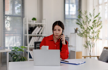 Obraz premium A professional woman engaged in a phone conversation while working on her laptop in a modern office space filled with natural light and greenery, showcasing productivity and communication.