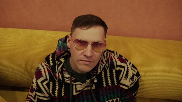 Man wearing stylish yellow sunglasses and patterned poncho sitting on yellow sofa. Portrait of relaxed male resting at home in cozy indoor environment. Lifestyle, fashion and leisure concept