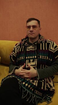 Man wearing stylish yellow sunglasses and patterned poncho sitting on yellow sofa. Portrait of relaxed male resting at home in cozy indoor environment. Lifestyle, fashion and leisure concept