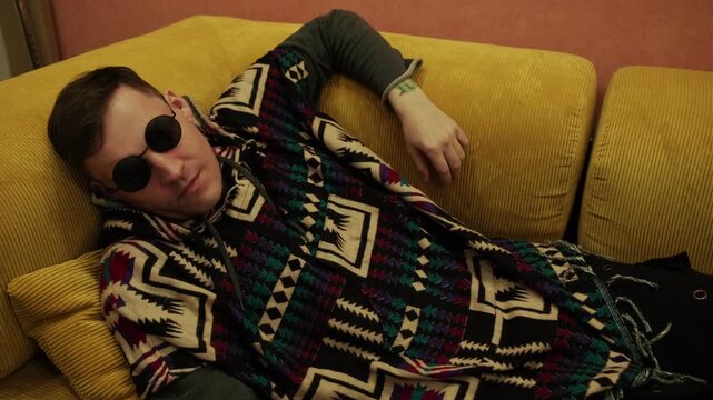 Man wearing ethnic hoodie and round sunglasses resting on couch. Hipster relaxing at home inside yellow interior. Young male in stylish patterned clothing resting on sofa hobby leisure.