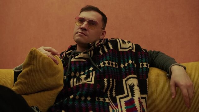Man wearing heart-shaped sunglasses and patterned hoodie sitting on yellow sofa. Stylish portrait of trendy male resting in modern lounge interior. Fashionable youth lifestyle concept.