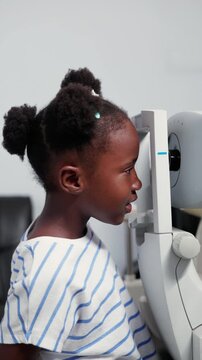 Vertical video, Young African girl child receiving an ocular examination using autorefractor machine in a clinic