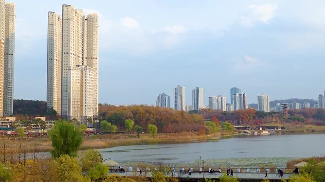 Aerial zoom out view of Gwanggyo Lake Park in autumn with colorful fall foliage, calm lake, pedestrian bridge and modern residential skyscrapers in Suwon Korea