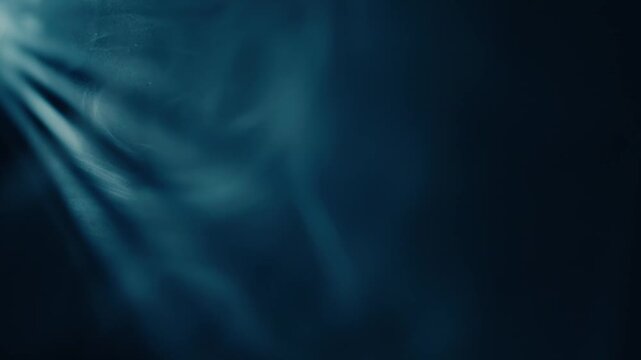 Intense blue rays of light shine through a dense, dark haze, illuminating floating particles. The shaft of light cuts through the shadows, creating a deep and mysterious visual effect.