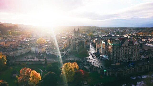 Beautiful golden hour aerial footage of the historic Bath Abbey and city center. The sun flares over the Roman-founded city, highlighting the classic honey-colored Georgian architecture and autumn tre