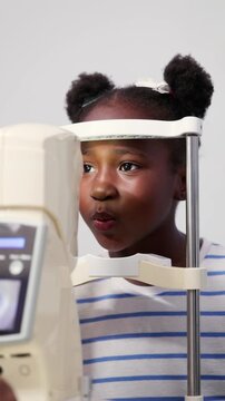 Vertical video, Young African girl focusing on a non-contact tonometer (NCT) device during an eye exam at an ophthalmologist's clinic