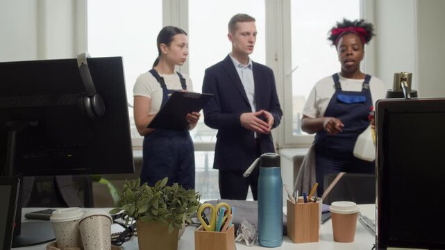 Tilt up shot of young Caucasian man talking to two female bicultural cleaners in uniforms while organizing office table cleanliness