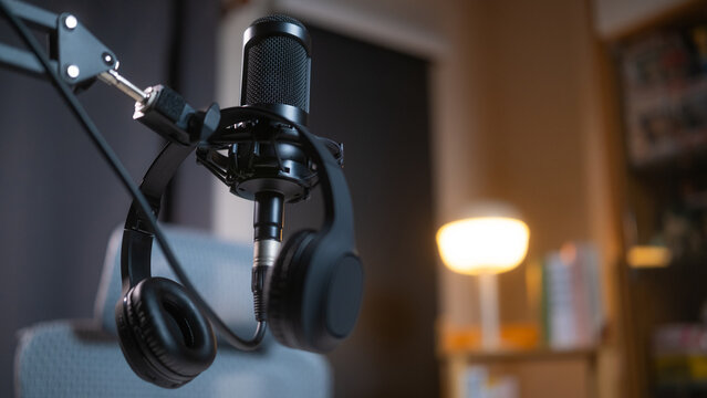 Close up of empty podcasting workspace with a professional broadcasting microphone and hanging headphones in sharp focus, set against a blurred background of a modern home recording studio.