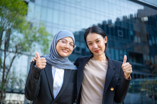 Portrait of two smiling diverse Asian businesswomen, one wearing hijab, happily thumbs up to the camera outdoors against a modern office building and green trees background.