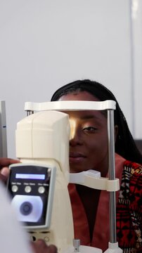 Vertical video, Young African woman having vision checked by optometrist using non-contact tonometer (NCT)