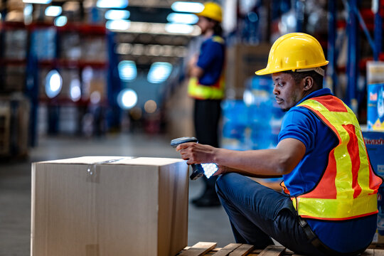 African American logistics worker checking cardboard box using barcode scanner and mobile device. Modern commercial freight shipping and supply chain operations.