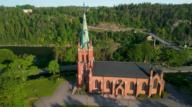 Trollh&auml;ttans Kyrka red brick Neo-Gothic church with copper spire revealing G&ouml;ta &auml;lv gorge granite cliffs and Olidan dam in golden evening light - aerial drone orbit