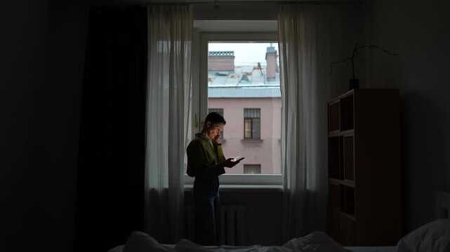 Upset shocked senior woman reading news or checking message on smartphone online, lady standing in dark bedroom in front of window with phone, elderly female with sad expression scrolls phone sighing.