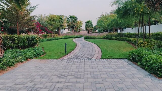 Walking path with lush green landscaping, featuring carpet grass, trees, palms and Washingtonias. Villas houses alongside greenery garden backyard. Interlocking stone pathway weaves through landscape.
