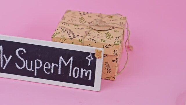 Hand present "My Super Mom" chalkboard sign front toa floral gift box on pink background