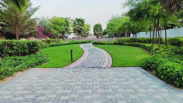 Walking path with lush green landscaping, featuring carpet grass, trees, palms and Washingtonias. Villas houses alongside greenery garden backyard. Interlocking stone pathway weaves through landscape.