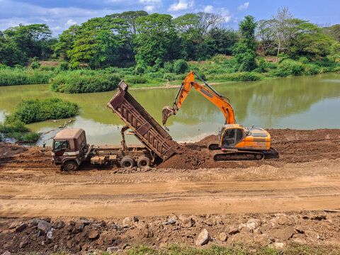 Construction work is taking place by the river; trucks are lifting their trailers to unload soil, and an orange backhoe is parked nearby, assisting in clearing the earth.