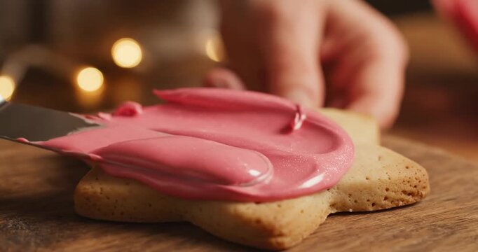 A hand delicately spreading vibrant pink icing on a star-shaped cookie, with festive lights in the background