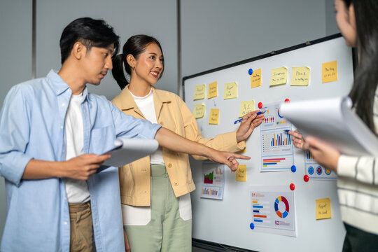 Asian happy business team discussing data charts and project plan on whiteboard in office, Young startup colleagues brainstorming marketing strategy, Corporate teamwork and presentation concept