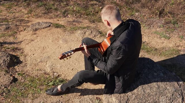 Blonde man playing string guitar outdoors in autumn forest. Concept of sound therapy, mental health and wellness rituals. Calmness tranquility audio-sensory practices. Aura farming energy holistic