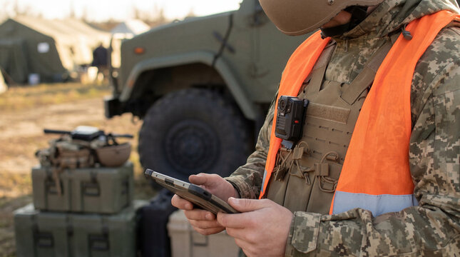 Soldier in camouflage uniform and orange safety vest, equipped with a body camera.