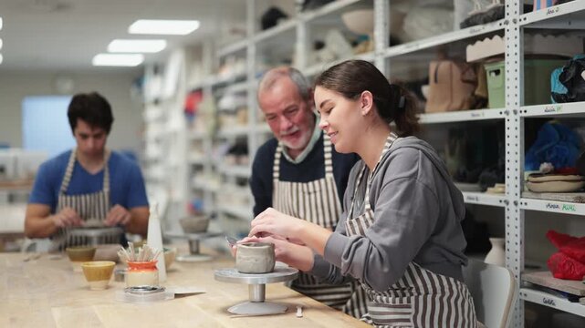 Eldery man helps and gives instructions to young woman sculpting clay at workbench in ceramic workshop