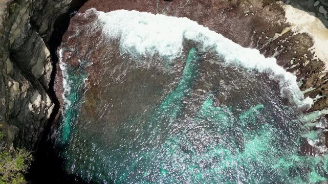 Aerial drone view of Broken Beach in Nusa Penida, Bali, Indonesia. Powerful ocean waves crash against the circular limestone cove, revealing turquoise water patterns over rocky reef formations