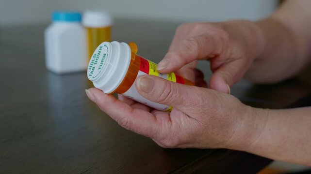 Close up of elderly hands holding and examining a prescription medication bottle with pharmacy label on a table.