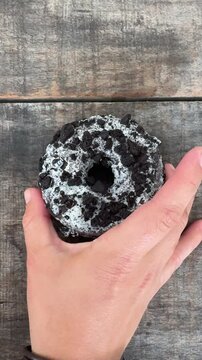 Hand picking up cookies and cream donut on wooden table. Casual UGC style food clip showing sweet dessert with crushed chocolate cookies and white glaze topping.