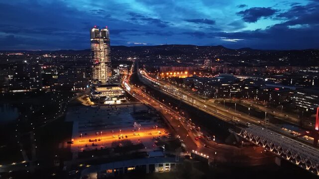 Aerial view of Budapest during blue hour, featuring the illuminated Mol Tower, nearby bridges, urban cityscape, and evening traffic against the scenic backdrop of the Buda Hills.