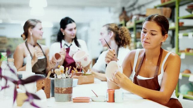 Interested young woman in pinafore drawing on handmade clay jar with paintbrush in art studio