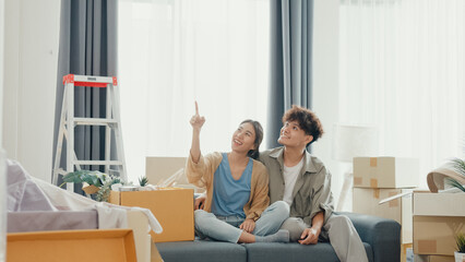 Young happy Asian couple sit on sofa couch use design thinking pointing wall get idea decoration...