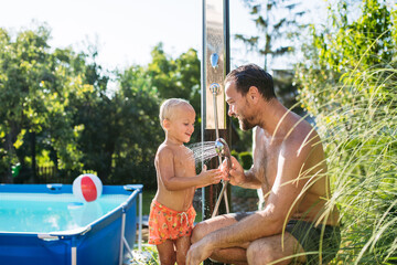 Son and father under shower by the pool on sunny summer day.