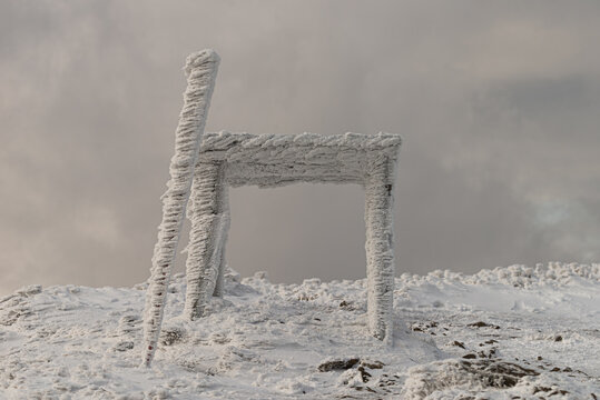 A wooden structure completely covered in thick rime ice and snow on Polonina Wetlinska, Bieszczady Mountains, Poland. Extreme winter weather conditions on a mountain peak under a cloudy sky.