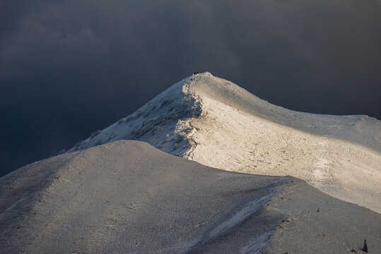 Bieszczady Mountains in winter, view of the snow-covered Polonina Carynska ridge under a dramatic dark sky, hikers on a mountain trail, Polish Carpathians landscape.