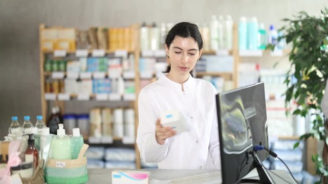 Near counter with computer is pharmacists workplace. Girl employee participates in online testing and learning, reads incoming emails. Electronic database of drugs. Colleague work in background
