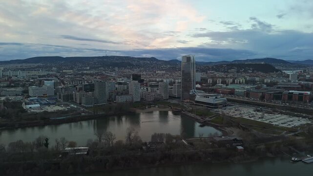 Drone view of Budapest at sunset, featuring the Mol Tower, modern architecture, and the Danube River, with a scenic urban skyline framed by the Buda Hills and Kopaszi G&aacute;t waterfront.