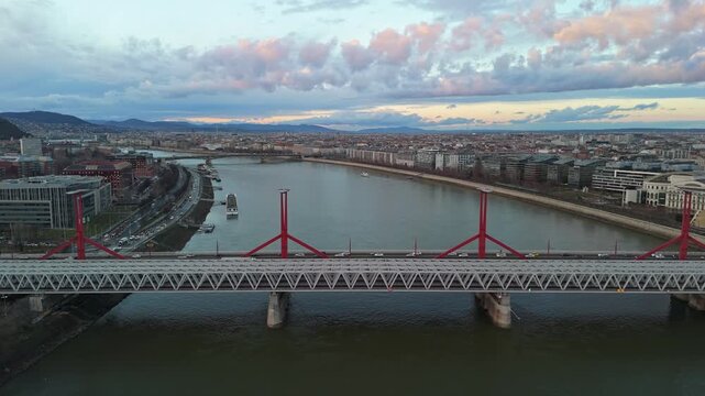 Sideways tracking aerial view of the R&aacute;k&oacute;czi Bridge over the Danube River in Budapest at sunset, showcasing urban architecture, traffic, and the scenic skyline of Hungary's capital city.