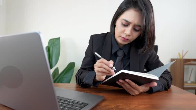 Closeup of upset working young Asian business woman sits on chair at workplace desk hold pen and diary making writing notes, doing online research network connection error problems.