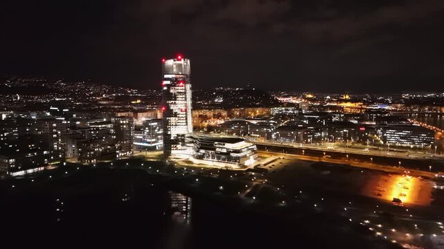 Orbital aerial view of Budapest at night, highlighting the illuminated Mol Tower, the Danube River, and the vibrant cityscape with traffic, modern architecture, and surrounding urban landmarks.