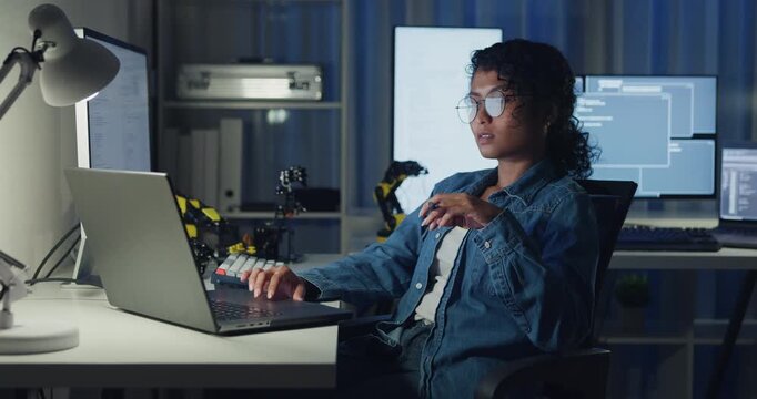 Young African American female programmer coding AI software on multiple screens in night office. Network security, cybersecurity, machine learning, working late, overtime, robotics automation concept.