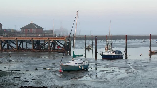 A panning shot reveals boats and sailboats moored in mudflats at low tide on a foggy morning in Gosport, England, with a pier and buildings in the background