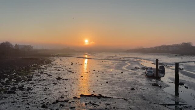 A wide shot captures a serene sunrise over the Gosport mudflats at low tide, with the sun reflecting on the wet mud and a bird flying across the hazy sky