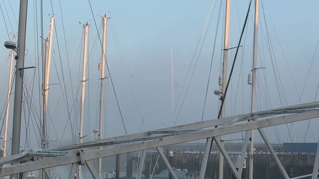 The Spinnaker Tower in Portsmouth, England, is partially obscured by fog, framed by the masts and rigging of boats in Gosport Marina on a cold morning