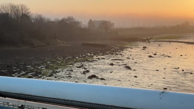 A pigeon takes flight from a railing, revealing a serene sunrise over the Gosport estuary with mudflats, small boats, and a misty golden sky