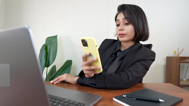 Close up of upset young Indian business woman sits on chair at workplace desk using smart phone network issue, low battery, wifi connection problem.