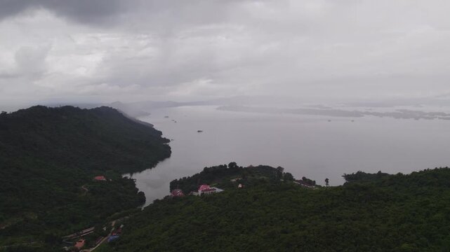 Very high altitude downward tilting and forward flying view of valley with the Nam Ngam lake and cloudy overcast sky in the background.