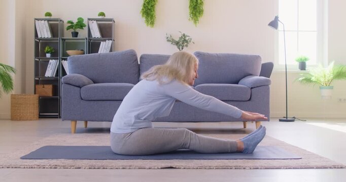 Senior elderly woman doing stretching exercises sitting on yoga mat at home. Mature female person exercising indoors. Healthy lifestyle, fitness and sport concept. 4k video. Slow motion video.
