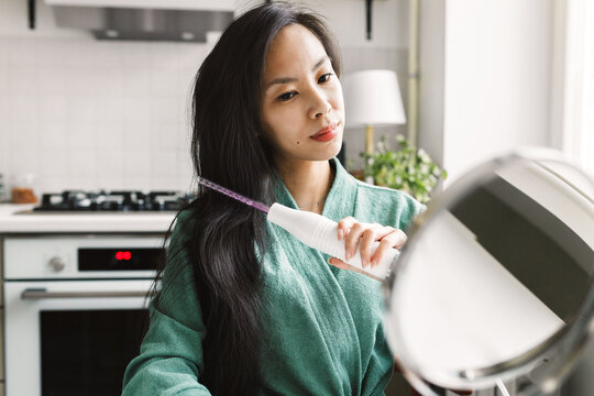 Selective focus on beautiful young asian woman brushing hair with darsonval in bathrobe, preventing and fighting with alopecia aretea at home, looking in round-shaped mirror standing on table