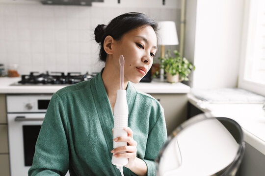 Selective focus on young asian girl woman in bathrobe using darsonval device for acne treatment, pore purification at kitchen table, looking in round-shaped mirror. Cosmetic spa procedures at home